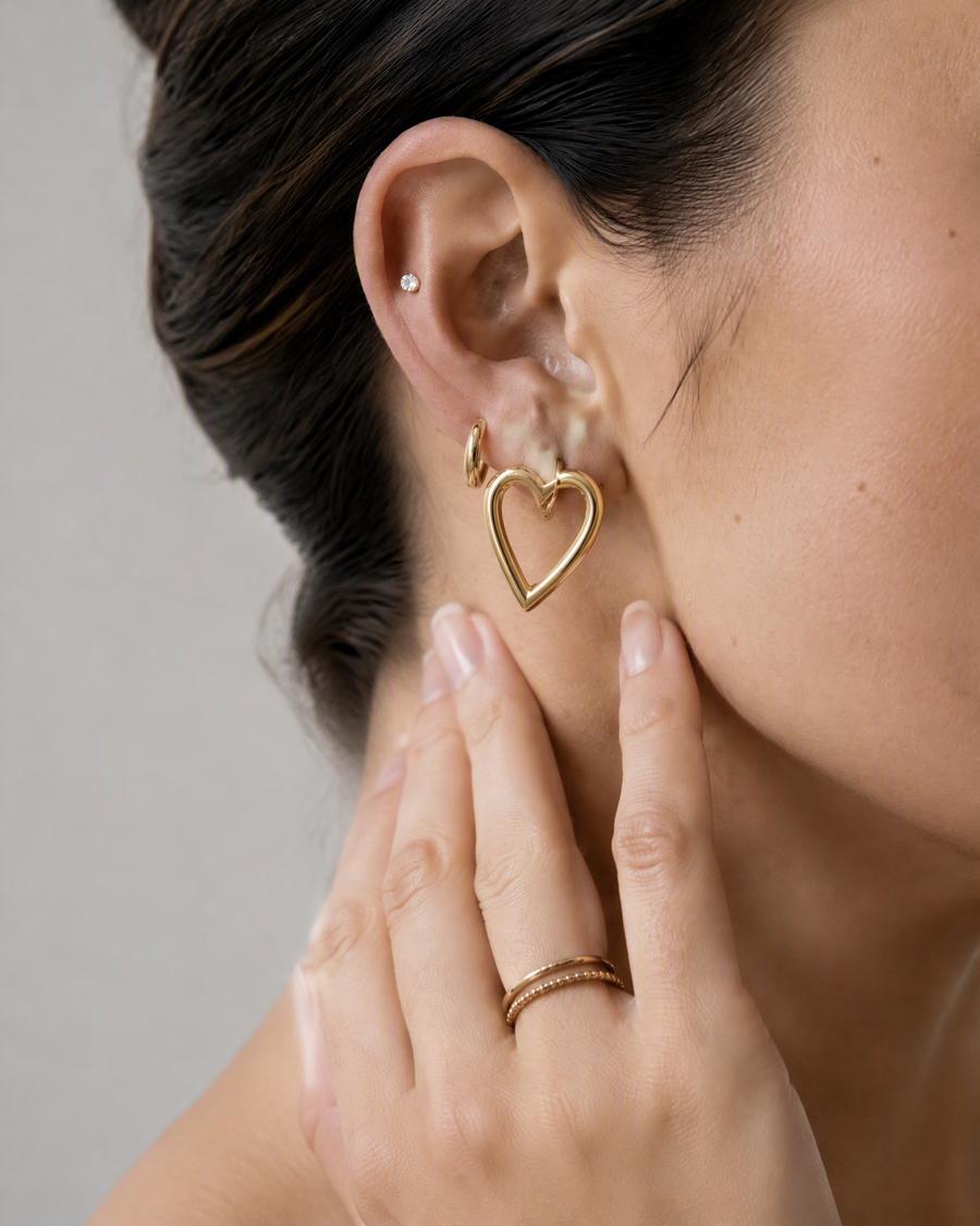 A close view of an ear wearing a large open heart drop, a small hoop and a tiny stud, with two slim rings on the hand below